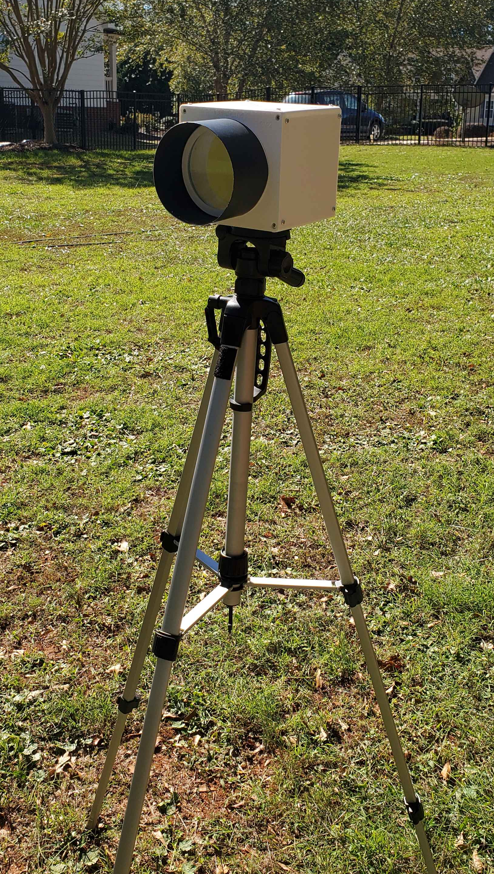 The 3D printed camera outside on a tripod. The camera is essentially a white cube with a lens on the front taking most of the space.sillo